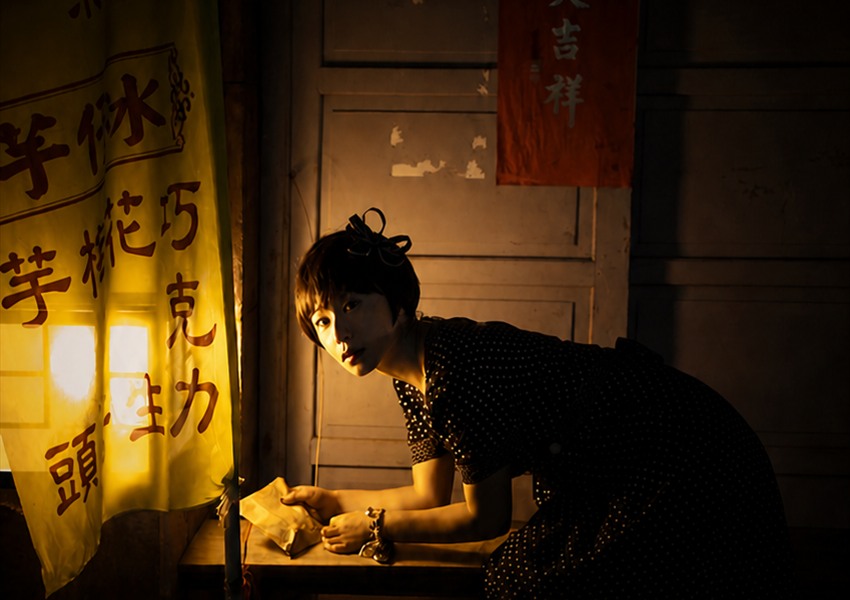 一位穿深色圓點洋裝的女子在暖黃燈光下俯身，背景是寫著漢字的布簾與紅色掛飾。